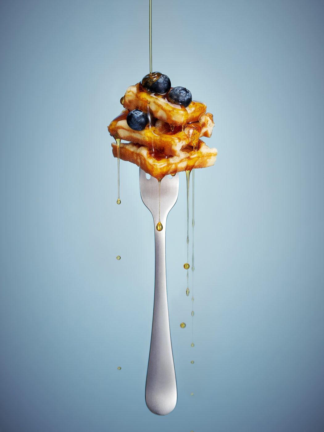 A floating fork pierces a piece of a waffle with a blueberry. Syrup pours from above.